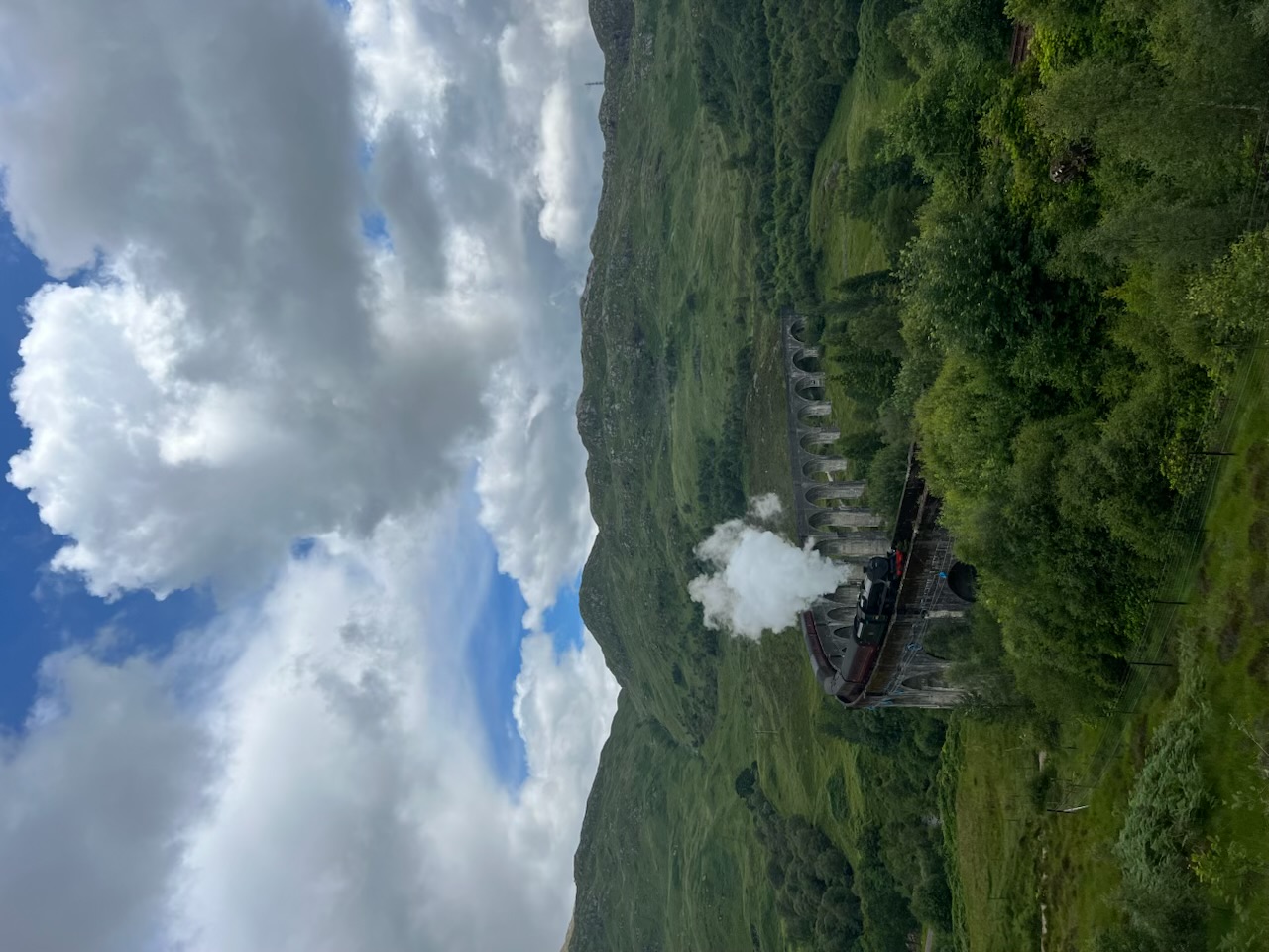 A steam train crossing a sweeping stone viaduct through green mountain valleys beneath a wide clouded sky